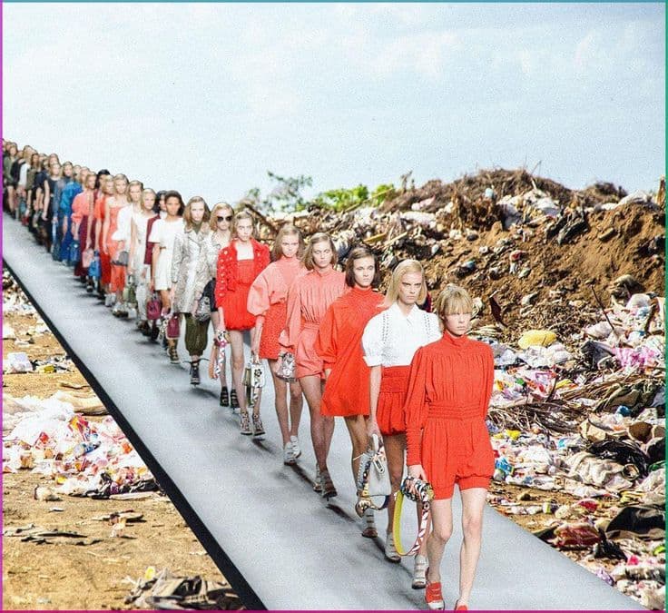 Runway show staged on a landfill, models in red and orange looks walking a path through waste